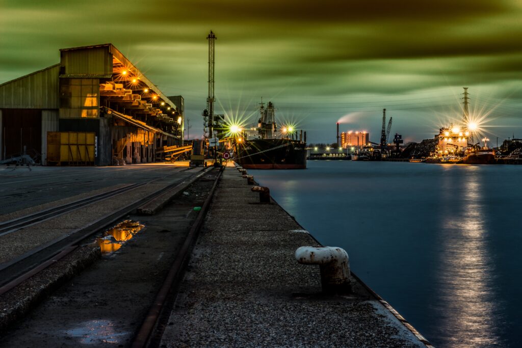 Uma vista noturna de um porto, com iluminação refletida na água e nuvens dramáticas ao fundo.