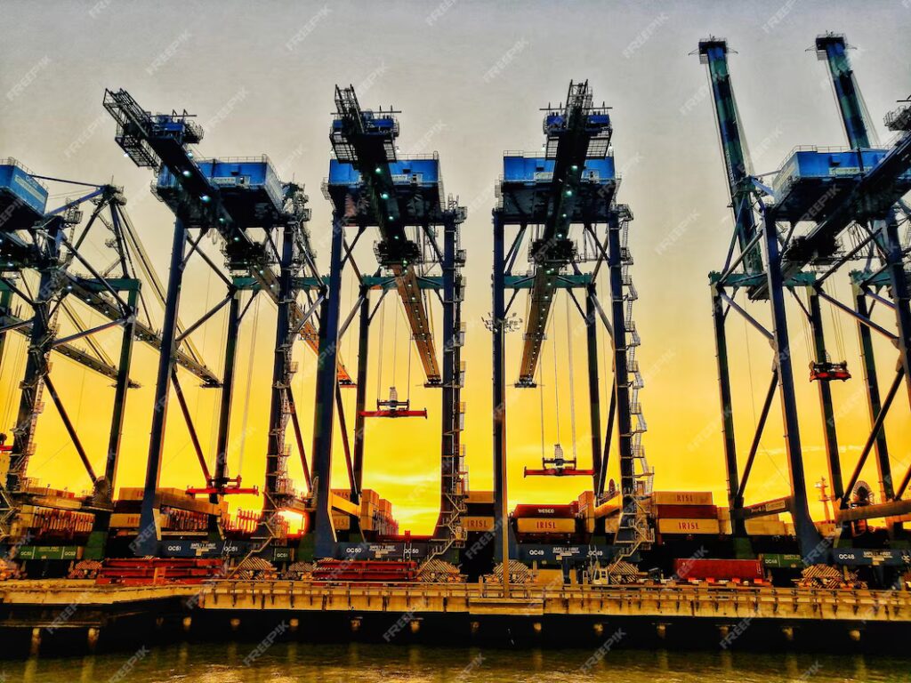 Guindastes em um porto ao pôr do sol, criando uma silhueta impressionante com contêineres.