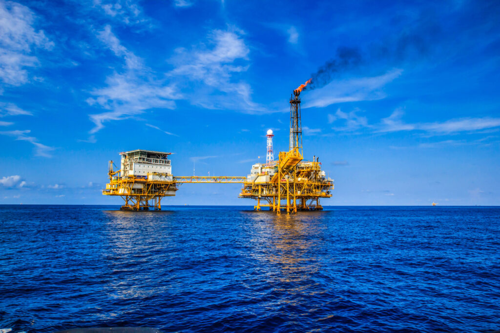 Uma plataforma de petróleo no mar sob um céu azul com nuvens e fumaça saindo de uma chaminé.